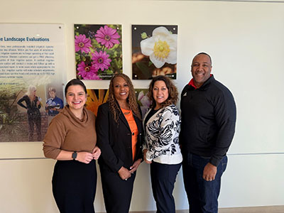 Four people stand smiling in front of a wall featuring two floral photos and a text display. The mood is friendly and professional.