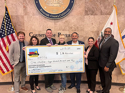 Seven people stand smiling and holding a large ceremonial check for $1.8 million. They are posed in front of the Riverside County seal and flags.