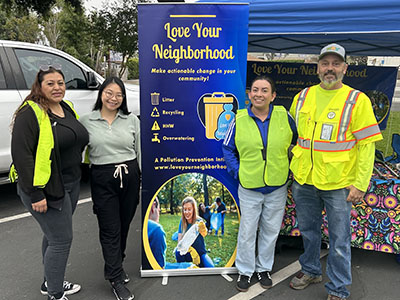 Four people wearing safety vests stand beside a "Love Your Neighborhood" banner at an outdoor event, promoting community recycling and environmental care.